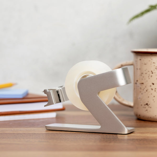 Side view of the silver tape dispenser showing its angled arm and weighted base on a desk
