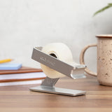 Silver tape dispenser on a desk beside sticky notes, pencils and a speckled mug