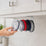 A hand taking a clear tumbler lid from a row of clear, red and navy lids hanging from the ELYPRO organizer under a white kitchen cabinet