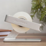 A new roll of tape being fitted into the black ELYPRO tape dispenser, the top arm lifted away and set back onto the weighted base, with the silver model shown first on a desk