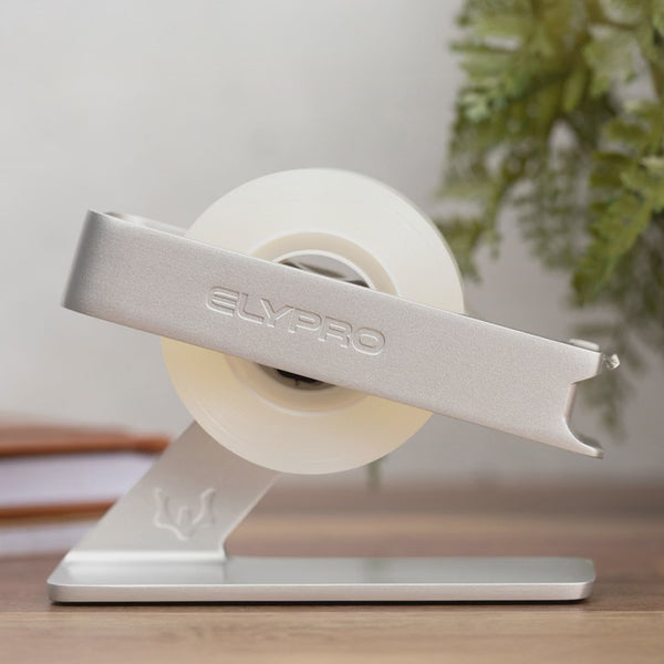 A new roll of tape being fitted into the black ELYPRO tape dispenser, the top arm lifted away and set back onto the weighted base, with the silver model shown first on a desk