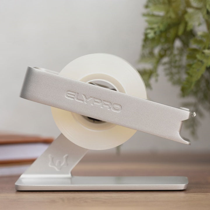 A new roll of tape being fitted into the black ELYPRO tape dispenser, the top arm lifted away and set back onto the weighted base, with the silver model shown first on a desk